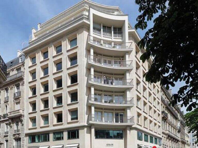 Un immeuble à plusieurs étages de l'avenue George V présente une façade incurvée avec de grandes fenêtres et plusieurs balcons. L'extérieur de couleur claire est complété par la verdure d'un arbre visible au premier plan à droite, encadrant partiellement l'image. - Paris Office Project