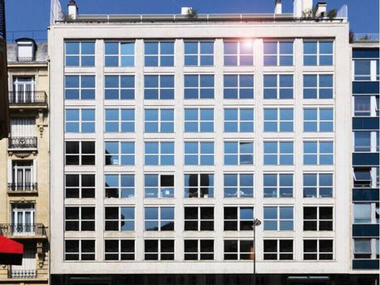 Un bâtiment moderne de plusieurs étages sur Pierre 1er de Serbie présente une grille de grandes fenêtres réfléchissantes sur la façade, présentant un design symétrique. Les bâtiments adjacents sont visibles de chaque côté, avec une lumière solaire reflétée dans le coin supérieur droit des fenêtres. - Paris Office Project