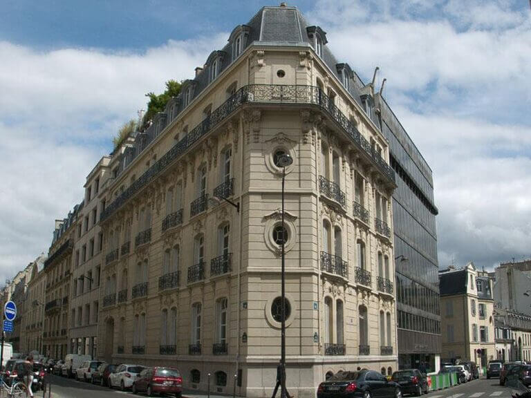 Un grand bâtiment orné se dresse au coin d'une rue animée de Paris, 75008. La structure présente des détails architecturaux complexes, notamment des fenêtres arrondies et des balcons en fer forgé. Les voitures sont garées le long du trottoir du 2 rue de la Baume, et le ciel est partiellement nuageux. - Paris Office Project