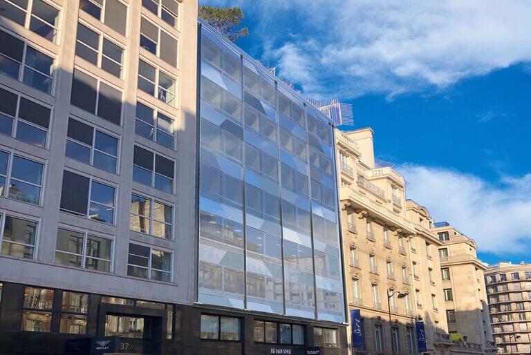 Une rue de la ville de Serbie présente un mélange de bâtiments modernes et traditionnels. Le bâtiment central présente une façade en verre reflétant le ciel bleu, tandis que d'autres structures sont en pierre avec des fenêtres rectangulaires. Le ciel est clair avec des nuages blancs épars, ajoutant à la scène pittoresque. - Paris Office Project
